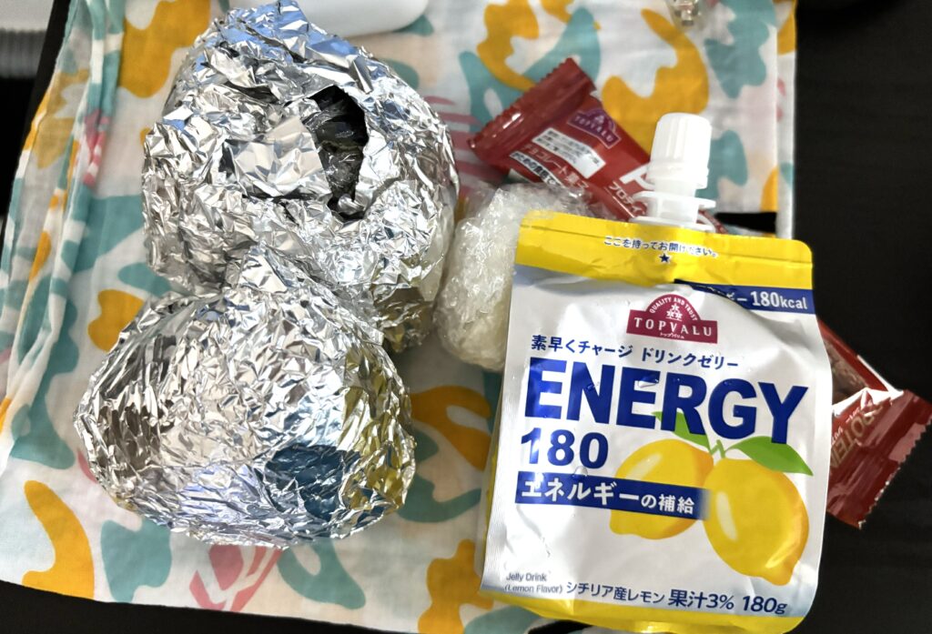 カラフルな布の上に置かれた補給食セット。アルミホイルに包まれたおにぎりが2つ、レモン味の「TOPVALU ENERGY 180」ゼリー飲料、そして小さなチョコレート菓子がある。