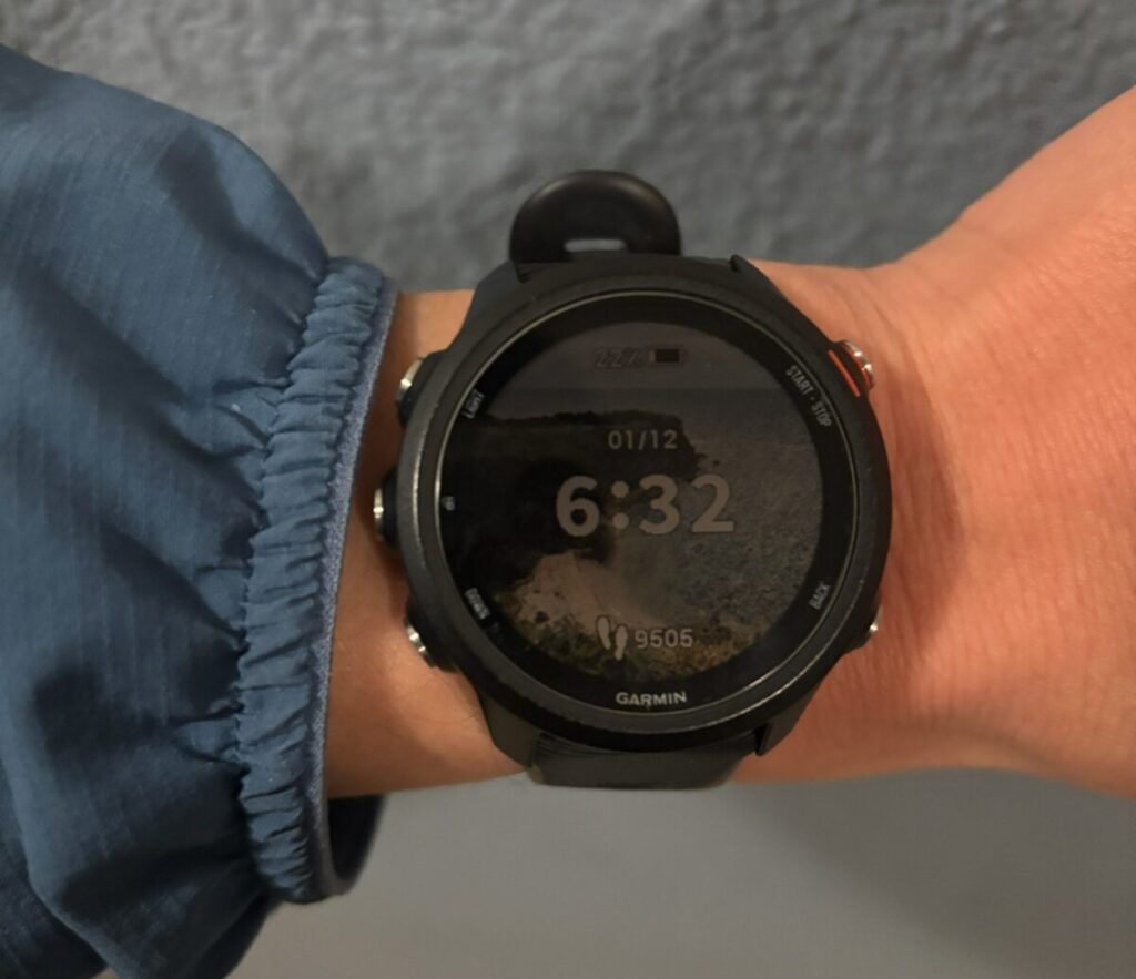 手首に装着されたGARMIN製の黒いスマートウォッチのクローズアップ。画面には日付「01/12」と時刻「6:32」、歩数「9505」が表示され、背景には風景写真が設定されている。手首には青いウェアの袖が見える。