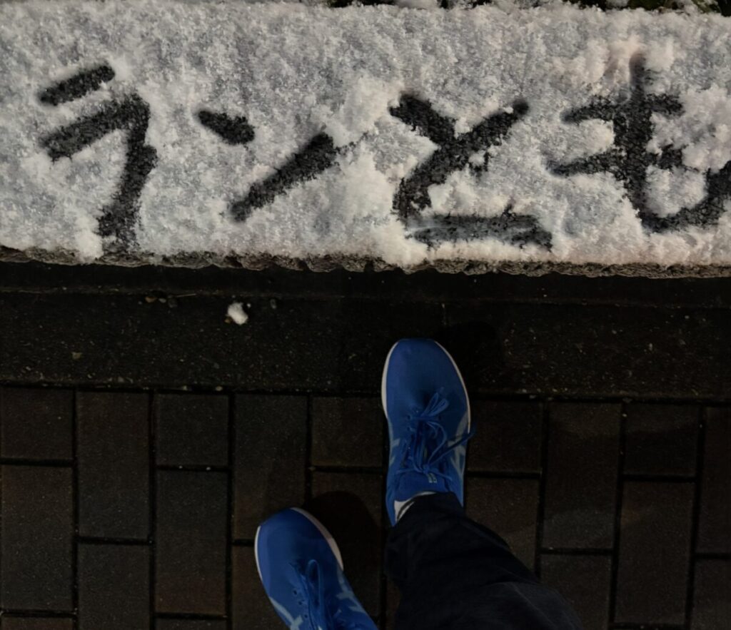雪が積もった地面に、手で書かれたと思われる日本語の「ランとも」という文字が黒く浮き上がっている。その下には、暗い舗装された路面に、鮮やかな青色のランニングシューズを履いた足元が写っている。全体として、夜間または薄暗い時間帯の屋外の情景を上から見下ろした構図。
