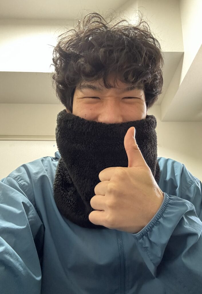青いフード付きジャケットを着た筆者Tomoyaさんが、黒いフリース素材のネックウォーマーで顔の下半分を覆い、目を閉じて満面の笑顔で親指を立てている。