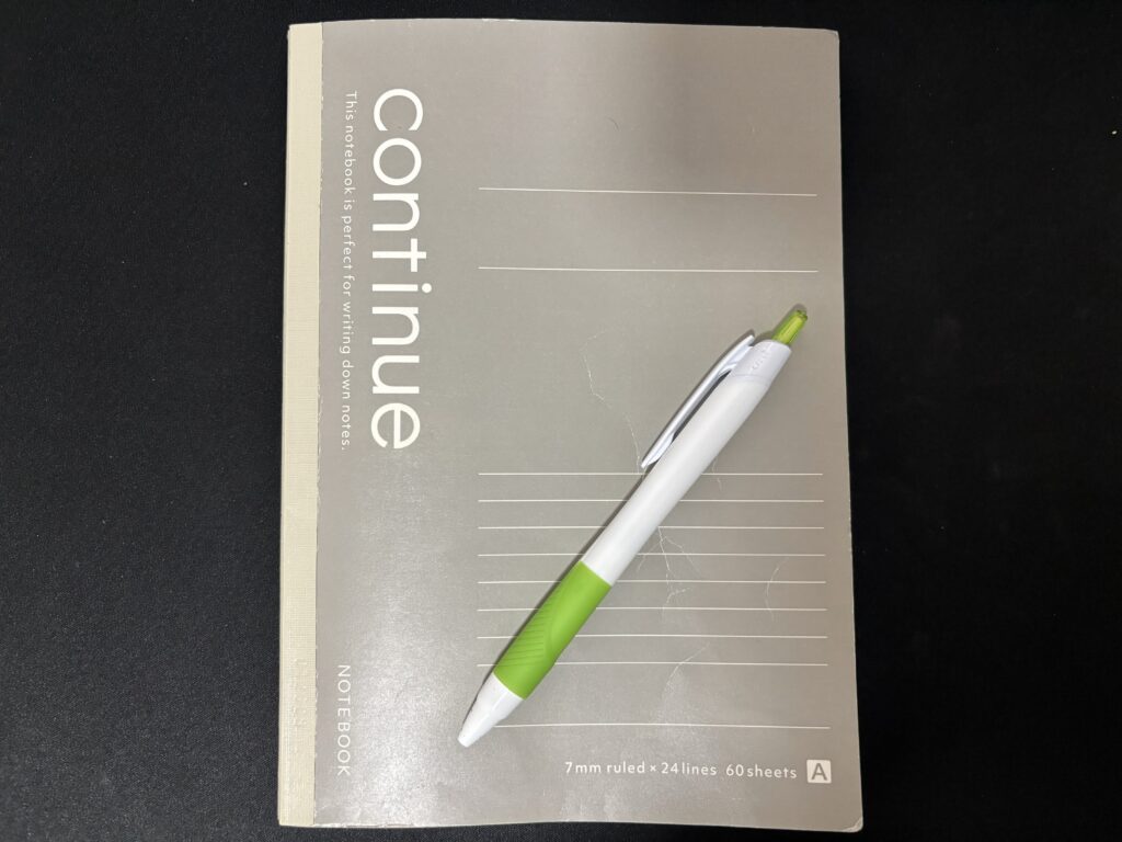「continue」と書かれたグレーのノートと、白と緑のボールペンが黒いテーブルに置かれている。ノートの表紙には「This notebook is perfect for writing down notes.」という英文と罫線の情報が記載されている。