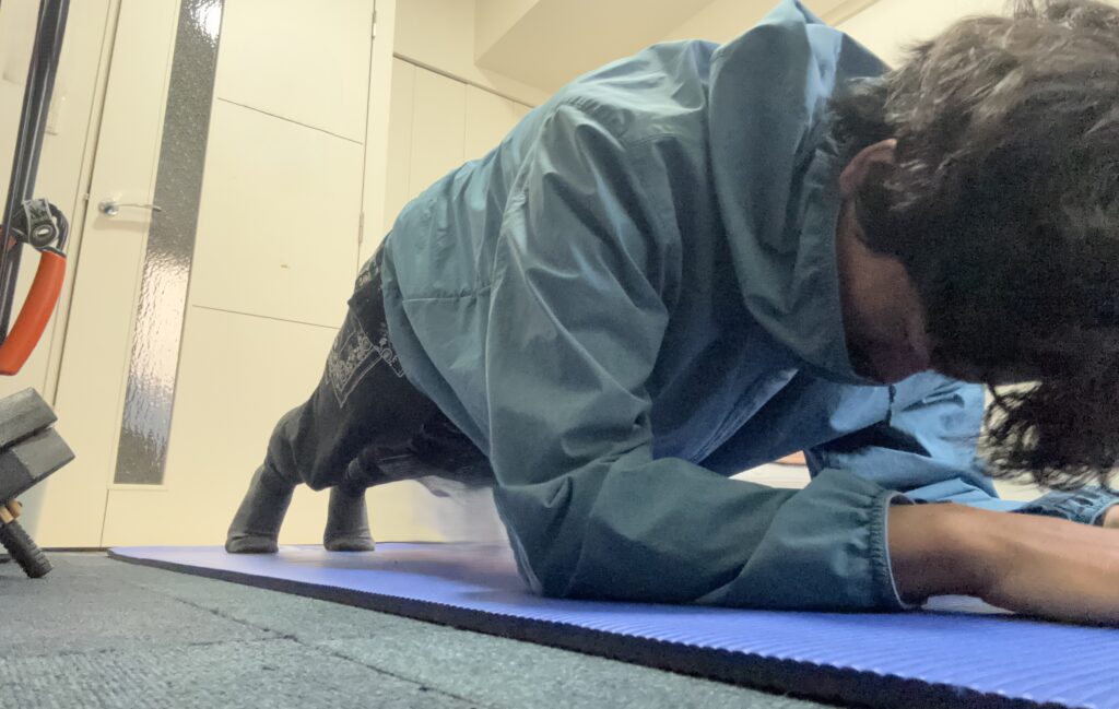 青みがかったフード付きジャケットと黒いズボンを着用した男性が、青いヨガマットの上でプランクをしている様子をローアングルから撮影。男性はマットに肘と前腕、床につま先をつけて体を支えている。