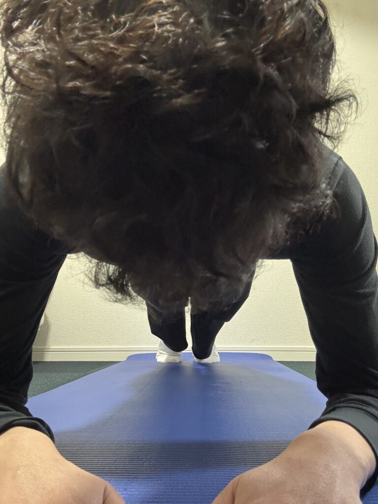 青いヨガマットの上で、黒い長袖シャツとズボンを着用した男性がプランクをしている様子を頭上から見下ろすアングルで撮影。男性の頭頂部と、マットに置かれた両腕、遠くには足が見える。