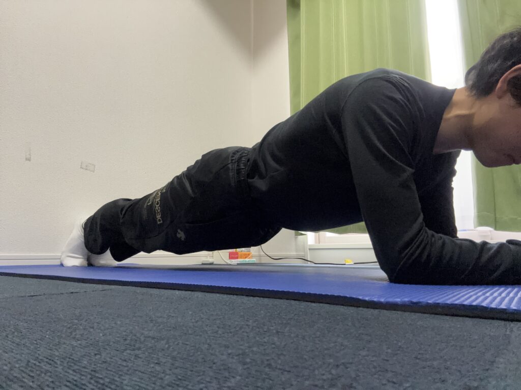 黒い長袖シャツとズボンを着用し、白い靴下を履いた男性が青いヨガマットの上でプランクをしている様子を側面から撮影。肘とつま先で体を支え、背中が一直線になる正しいフォームが保たれている。
