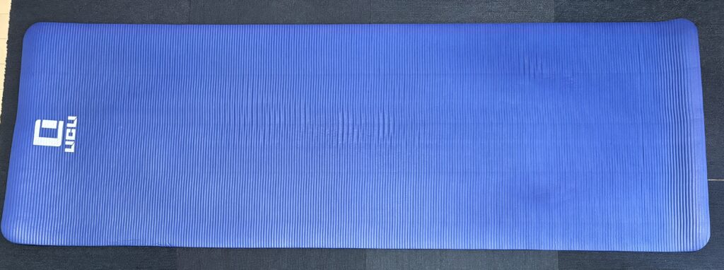 床に広げられた青いヨガマットを上から見た写真。マットには縦方向の溝があり、左上には「UGU」と書かれた白いロゴマークが見える。マットの下は濃い灰色のカーペット。現在、筆者(Tomoya)が使っているヨガマットの写真。長さ約183cm。