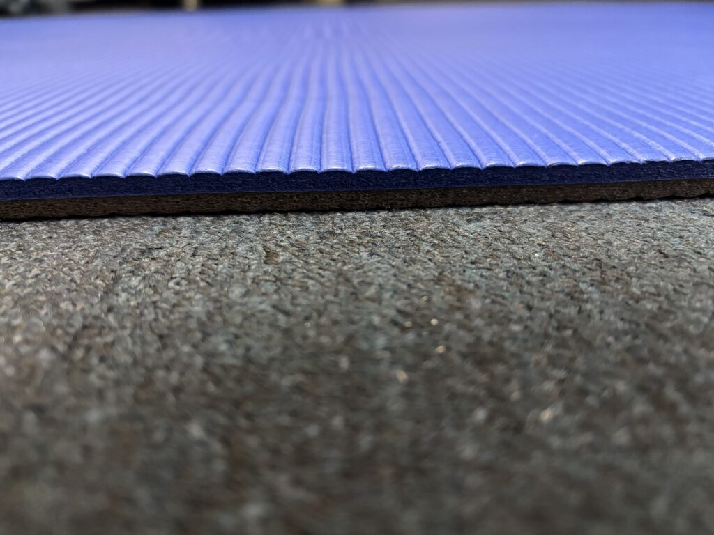 濃い灰色のカーペットの上に敷かれた青いヨガマットを、横から低いアングルで撮影した写真。マットの厚みと表面の縦の溝の質感がクローズアップされている。厚み約10mm。