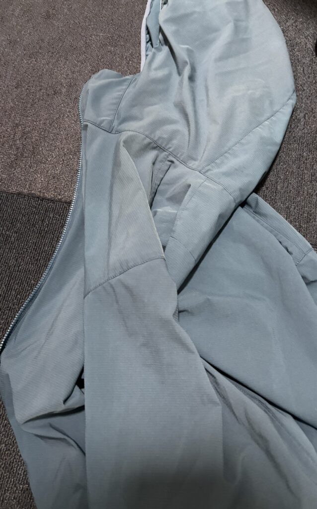 濃い灰色のカーペットの上に広げられた青みがかったグレーのフード付きジャケット。ジッパーは開いており、袖やフードの部分に皺が見える。
