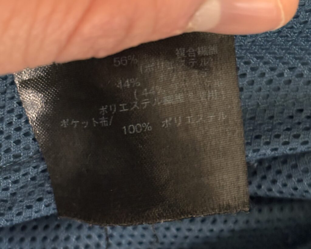 指でつまんで示された衣類の黒いタグ。タグには「56% 複合繊維 (ポリエステル) 44% ポリエステル繊維使用 ポケット布/ 100% ポリエステル」と日本語で素材の組成が表示されている。背景には青いメッシュ素材が見える。