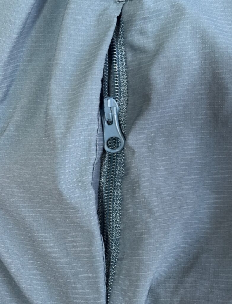 青みがかったグレーの生地に縦の線が入ったジャケットのジッパー部分のクローズアップ写真。ジッパーは半分ほど閉じられており、引き手が見えている。