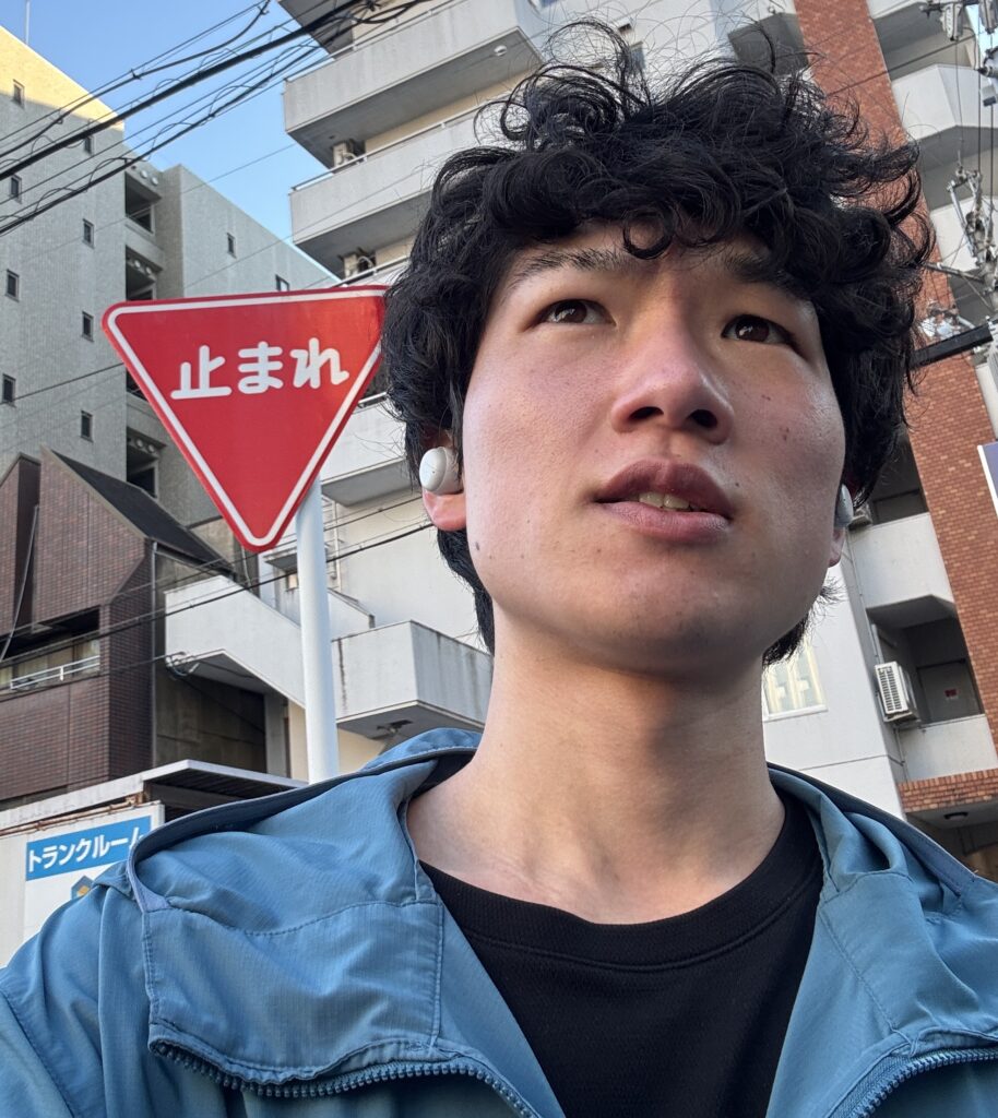 青いジャケットを着た筆者Tomoyaさんが、屋外で上を見上げて考え込んでいるような表情をしている。背景には建物と「止まれ」の赤い交通標識が見える。Tomoyaさんは白いワイヤレスイヤホンを装着している。