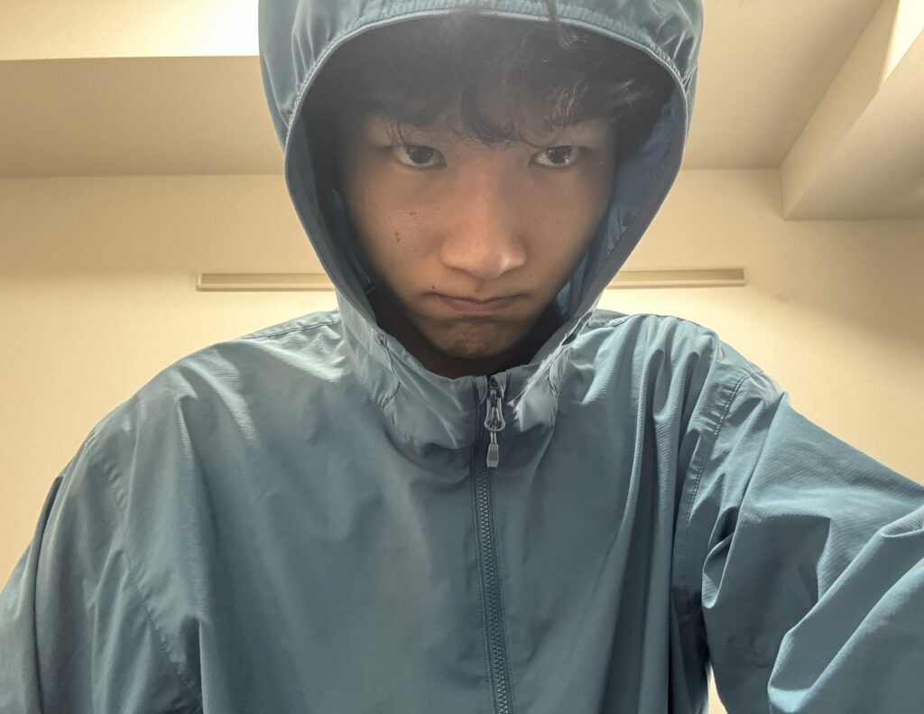 青いフード付きジャケットを着用した若い男性(筆者Tomoya)が、カメラに向かってまっすぐ真剣な表情を浮かべ、少し口をすぼめているクローズアップ写真。