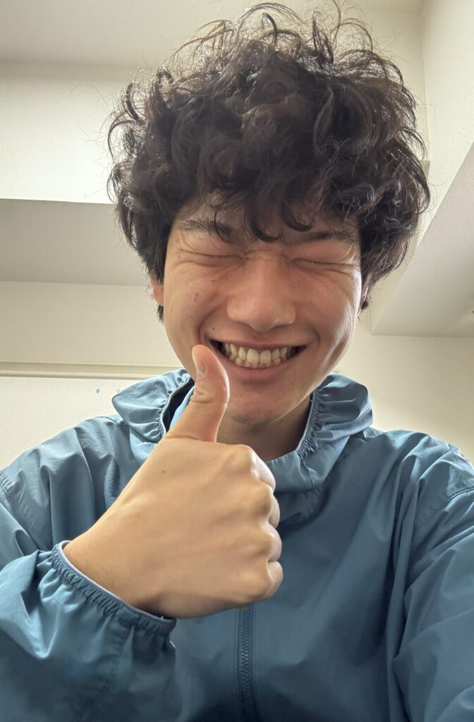 青灰色のフード付きジャケットを着た若い男性(筆者Tomoya)がカメラに向かって親指を立てて良いねを示すジェスチャーをしている。