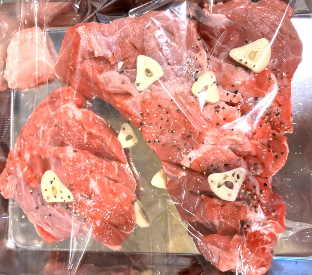 銀色のトレーの上でラップに包まれた生の牛肉の塊が2つ。肉の表面にはスライスされたニンニク片と黒胡椒が散らされており、調理前の下準備がされている様子がうかがえる。
