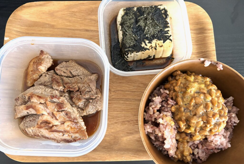 木のトレイに置かれた3品からなる食事。左にカットされた調理済みの牛肉が容器に入っており、右上に刻み海苔が乗った豆腐の容器、右下には雑穀米の上に納豆が乗った茶碗が配置されている。