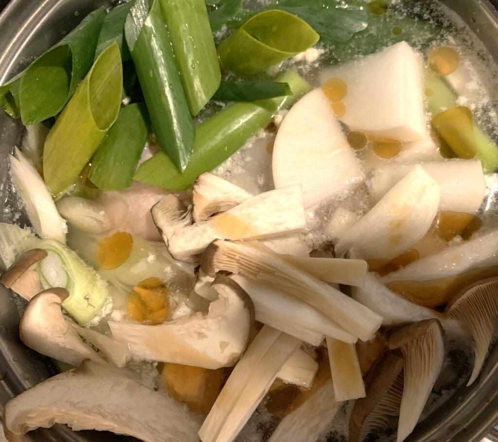ステンレス製の鍋で煮込まれている野菜とキノコの写真。厚めに切られた長ネギ、大根、エリンギやしめじのキノコ類が透明なスープに浸っており、少量の黄色い油滴が浮いている。