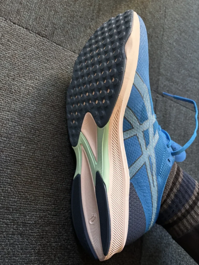 灰色の布地の上で、青いランニングシューズの裏側と側面が見えるように置かれている。アウトソールは黒、水色、クリーム色で構成され、多くの丸い突起が特徴的。靴下を履いた足首が少し写り込んでいる。