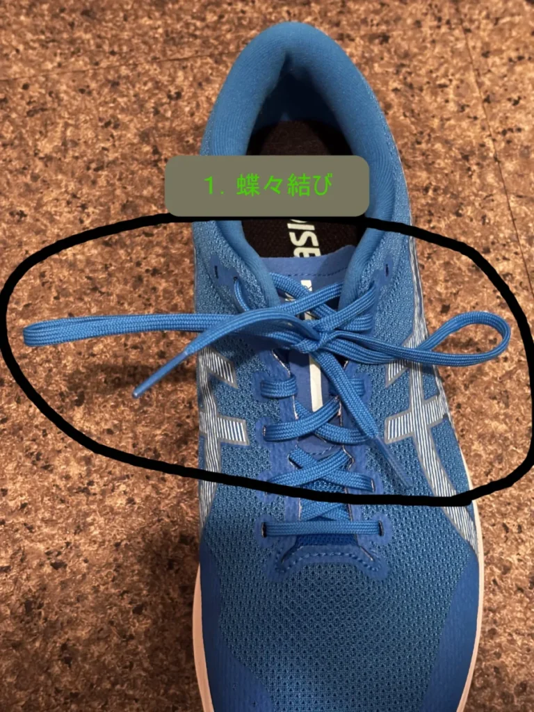 青いランニングシューズの靴紐が一般的な蝶々結びで結ばれている様子を上から撮影した写真。結ばれた靴紐の部分が黒い円で囲まれ、その下に「1. 蝶々結び」という手順を示すテキストがある。