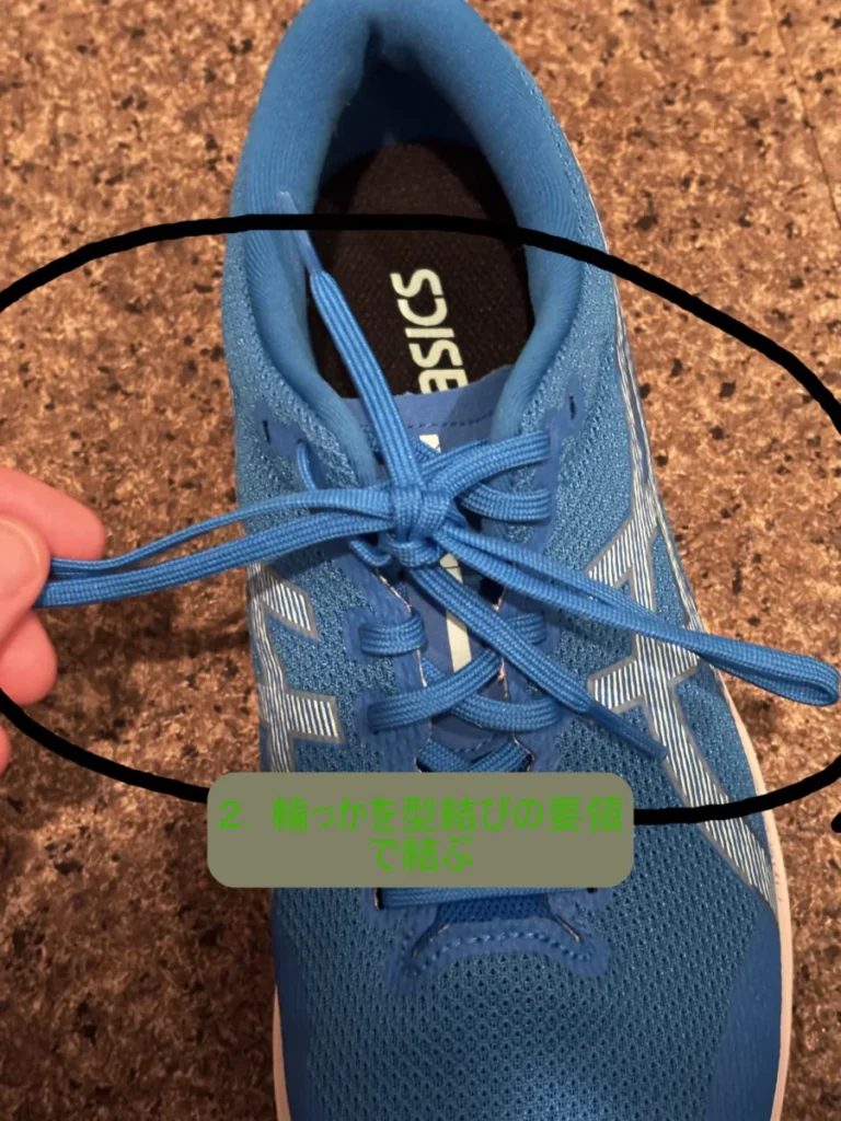 青いランニングシューズの靴紐を結ぶ途中を上から撮影した写真。すでに蝶々結びの片方の輪が作られており、もう一方の紐でその輪を結ぼうとしている段階が黒い円で囲まれている。その下に「2. 輪っかを型結びの要領で結ぶ」という手順を示すテキストがある。