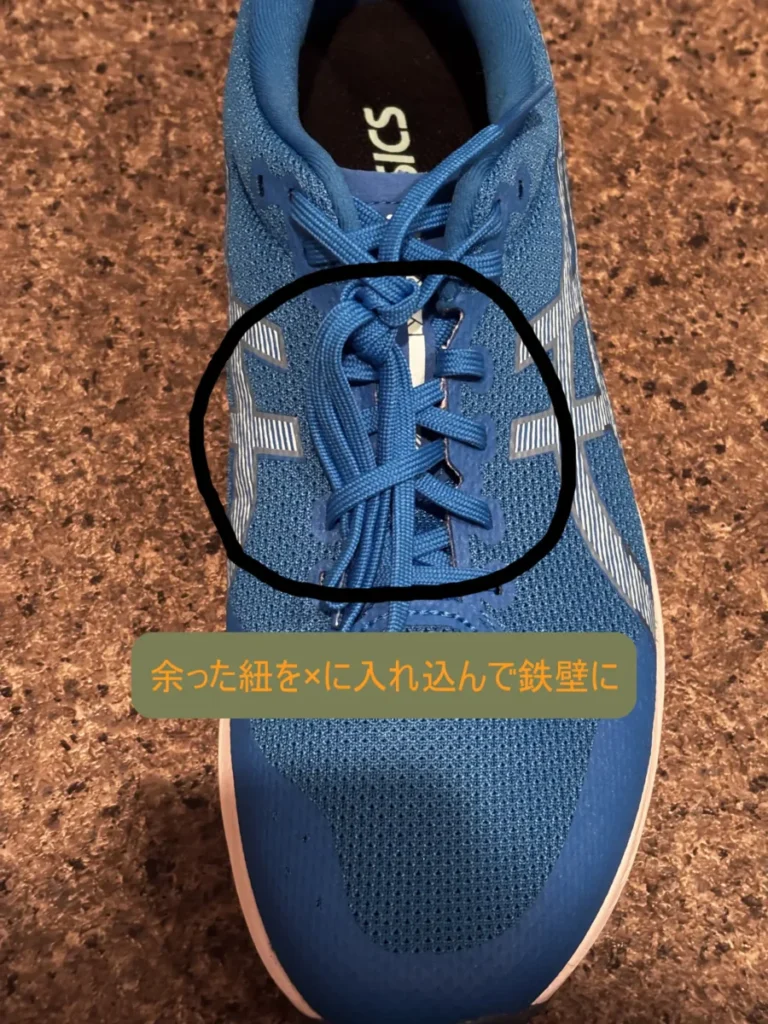 青いランニングシューズの靴紐が結ばれた後、余った紐がシューズの甲の部分に交差するように押し込まれている状態を上から撮影した写真。押し込まれた紐の部分が黒い円で囲まれ、その下に「余った紐を×に入れ込んで鉄壁に」というテキストがある。