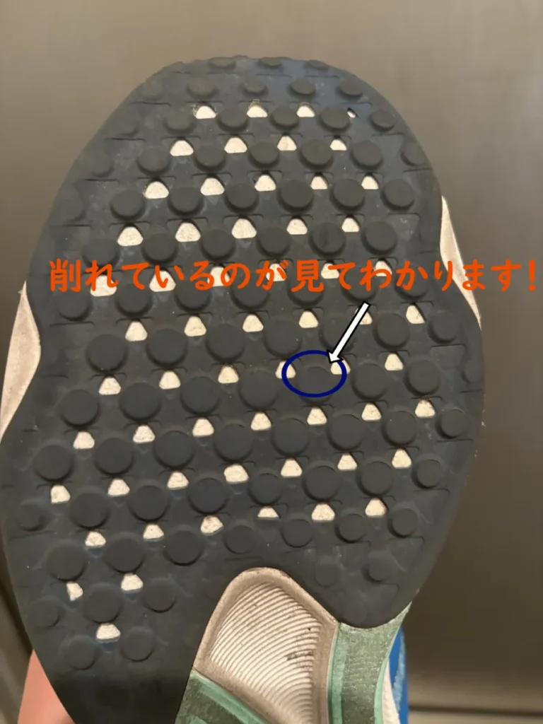 ランニングシューズの靴底をクローズアップした写真。黒いゴムのソールには多くの丸い突起があり、中央付近の一点が青い丸で囲まれ、白い矢印で示されている。その部分が摩耗して削れているのがはっきりとわかる。上部に「削れているのが見ててわかります!」という日本語のテキストが書かれている。2026年1月12日の写真。1か月使った後のソール(グリップ)